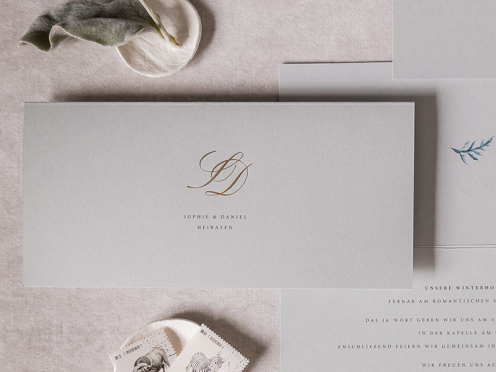 Erleben Sie elegante Einladungen zur Hochzeit mit Ihrem individuellen Monogramm in Goldprägung auf edlem grauem Papier. Der Briefumschlag, verziert mit einem goldenen Herz, ergänzt den exklusiven Look perfekt.