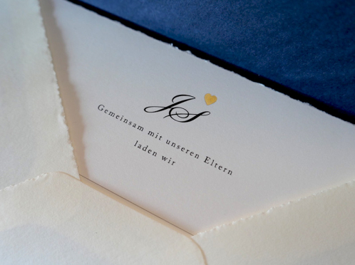 Diese exquisiten Einladungskarten auf cremeweißem Büttenpapier, veredelt mit mattgoldener Herzprägung, werden von einem blau gefütterten Umschlag ergänzt.