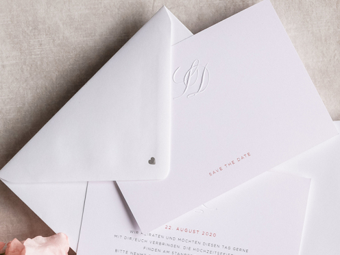 Mit persönlichem Monogramm geprägte Save-the-Dates, gedruckt auf weißem GMUND Papier. Ein silbernes Herz schmückt die Rückseite des Briefumschlags.