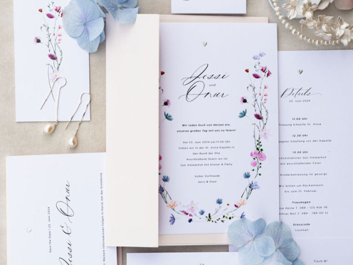Die Hochzeitseinladung vereint zarte pastellige Blumenranken, minimalistische Typografie und ein silbernes Herz als handgeprägtes Detail – elegant, romantisch und zeitlos.