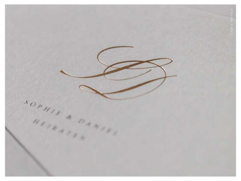 Erleben Sie elegante Einladungen zur Hochzeit mit Ihrem individuellen Monogramm in Goldprägung auf edlem grauem Papier. Der Briefumschlag, verziert mit einem goldenen Herz, ergänzt den exklusiven Look perfekt.